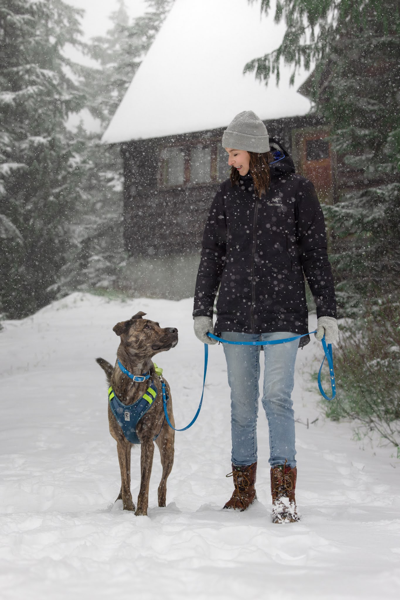 Person in winter clothing stands with a dog on a leash in a snowy landscape, with a wooden cabin in the background.